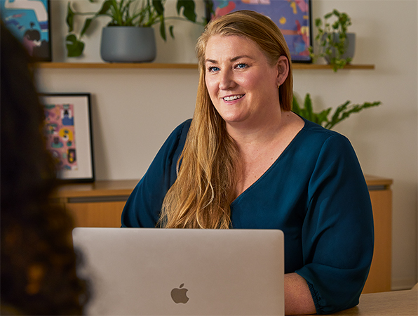 Une employée?Apple est assise à une table dans un espace collectif. Elle utilise un MacBook et discute avec une collègue.