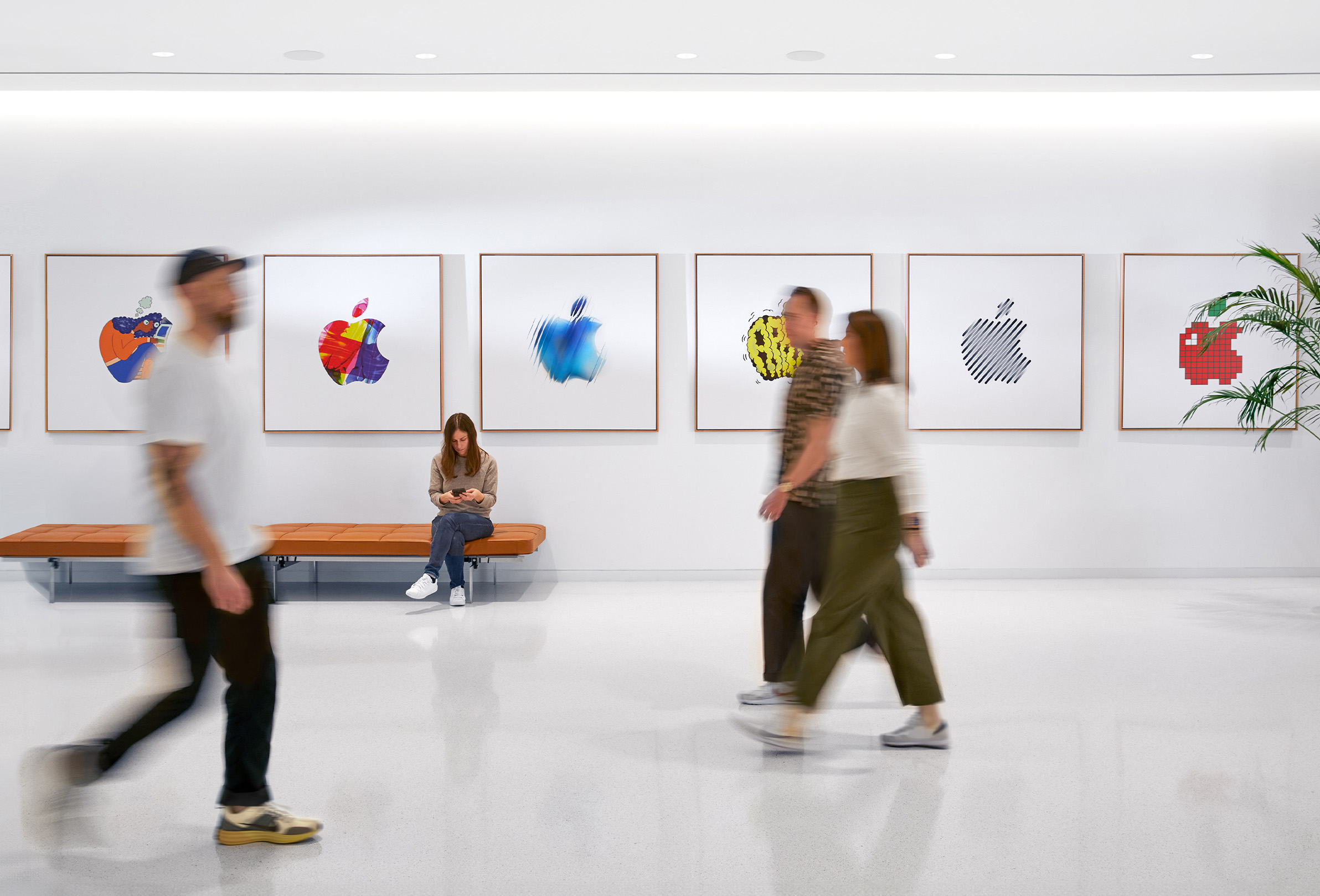 Trois membres de l’équipe Apple marchent devant une cloison décorée de logos?Apple colorés. Une quatrième employée est assise sur un?banc.