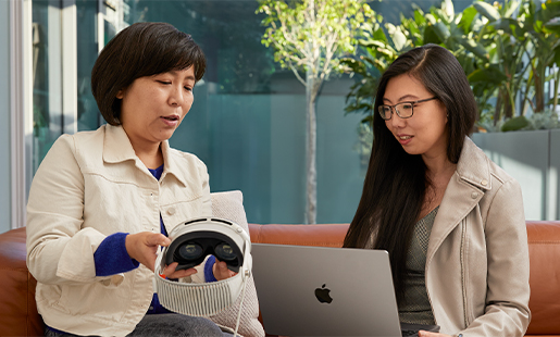 Jessica utilise un MacBook dans un espace intérieur, devant une baie vitrée qui donne sur une terrasse. Elle est assise avec une collègue?Apple qui lui montre l’Apple?Vision?Pro.