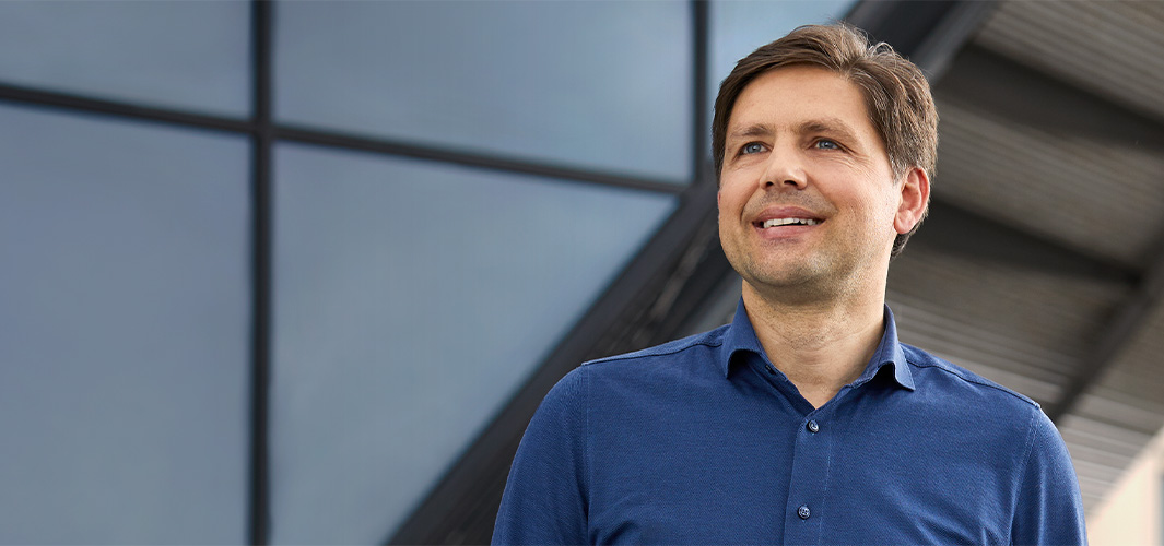 Greg stojí venku p?ed stěnou s?okenními tabulemi, dívá se doleva a?usmívá se.
