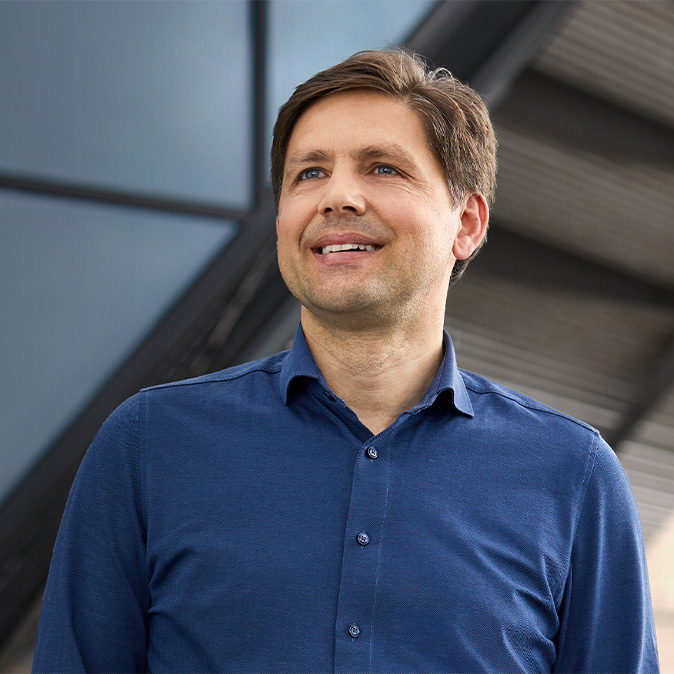 Greg stojí venku p?ed stěnou s?okenními tabulemi, dívá se doleva a?usmívá se.