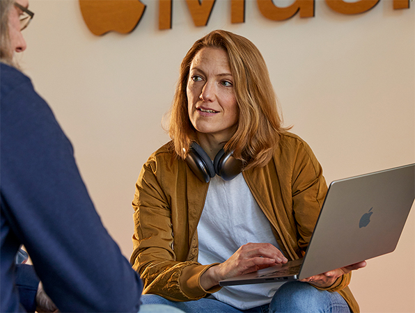 Two Apple Sales and Business Development team members in conversation, one holding a MacBook and sitting in front of a wall with the Apple?Music logo on it.