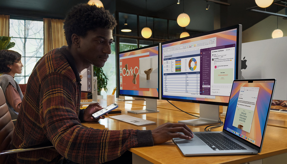 Une personne dans un espace de bureau utilise un MacBook Air ouvert connecté à deux écrans externes et à un Magic Keyboard, en tenant un iPhone dans l’autre main