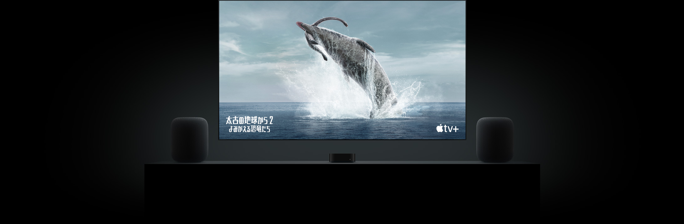 フラットテレビの大きなスクリーン上に「太古の地球から?よみがえる恐竜たち?」の恐竜の鮮明な映像が映し出されている。リビングルームのキャビネットの上に置かれた2臺(tái)のHomePodスピーカー。その間にApple TVが配置され、その上にテレビが掛けられている