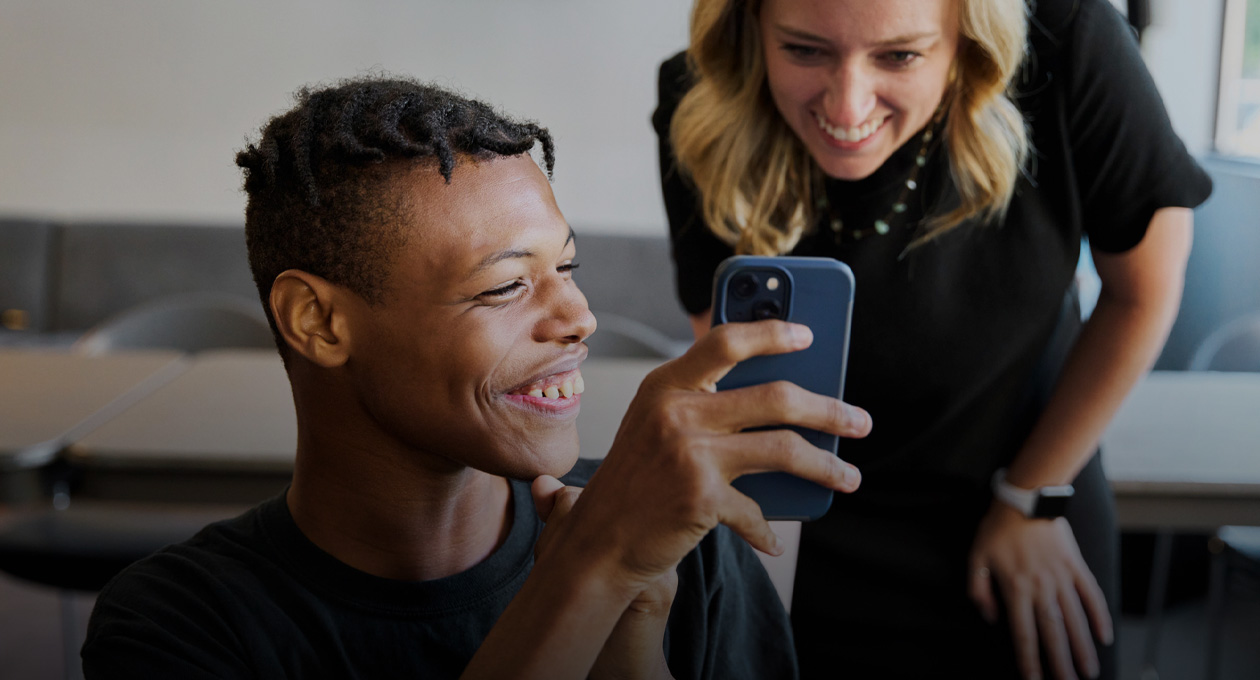 Foto de un joven con baja visión que sonríe y muestra la pantalla de su iPhone a una mujer, también sonriente, que está de pie detrás, a su izquierda