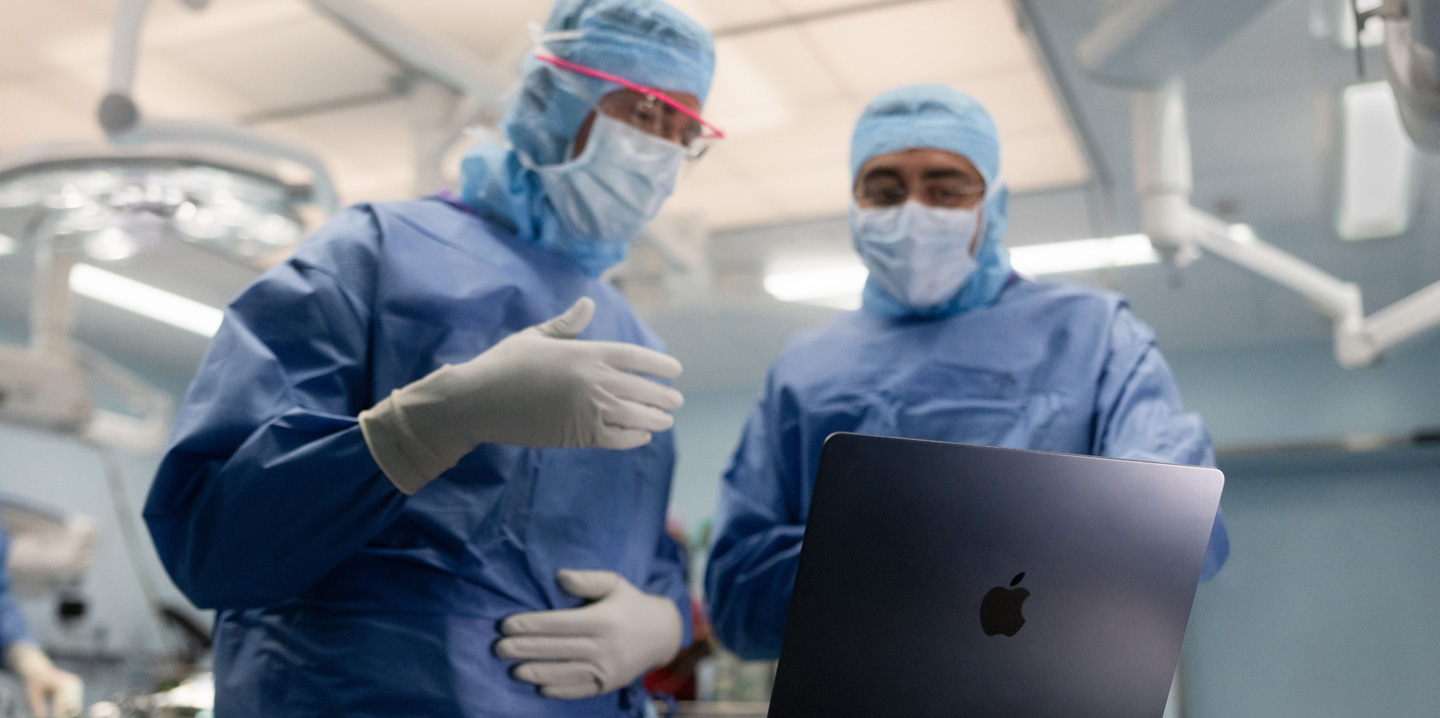 Due chirurghi in sala operatoria consultano un MacBook: uno fa un segno con la mano, mentre l’altro osserva